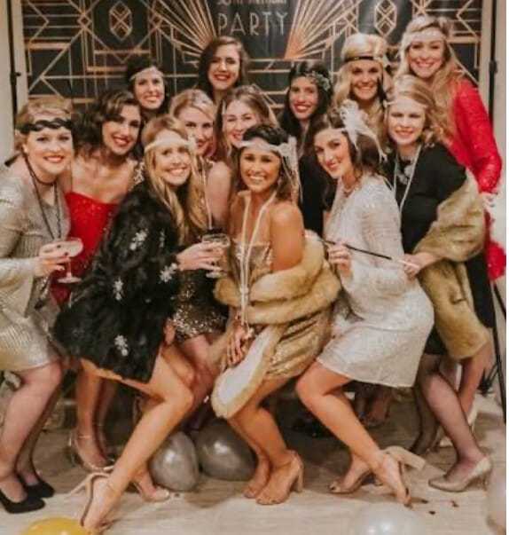 Group of women in 1920s-themed outfits at a festive gathering, smiling and holding drinks.