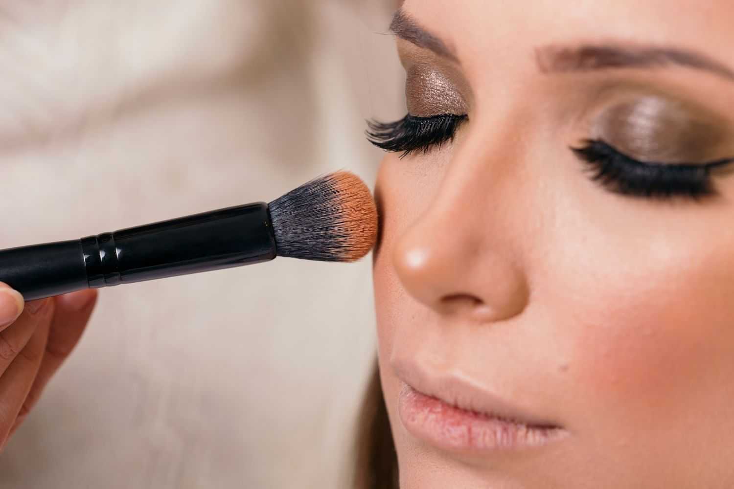 Makeup brush applying blush on woman's cheek with eyeshadow and false lashes.