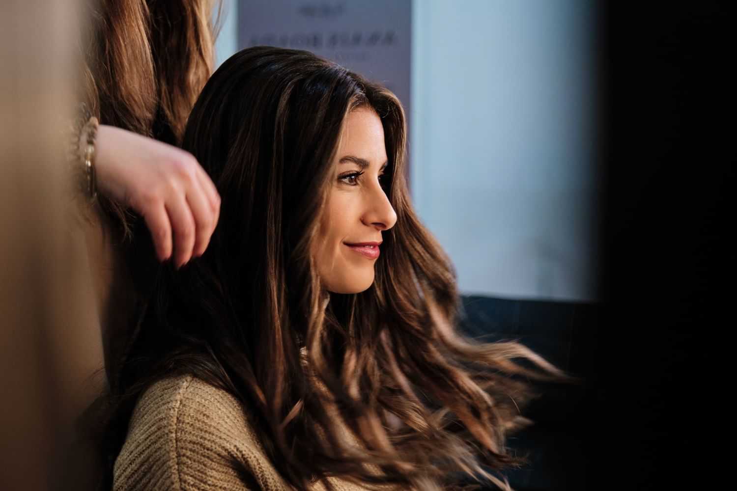 Hair stylist fixing woman's long, wavy brown hair inside a salon.