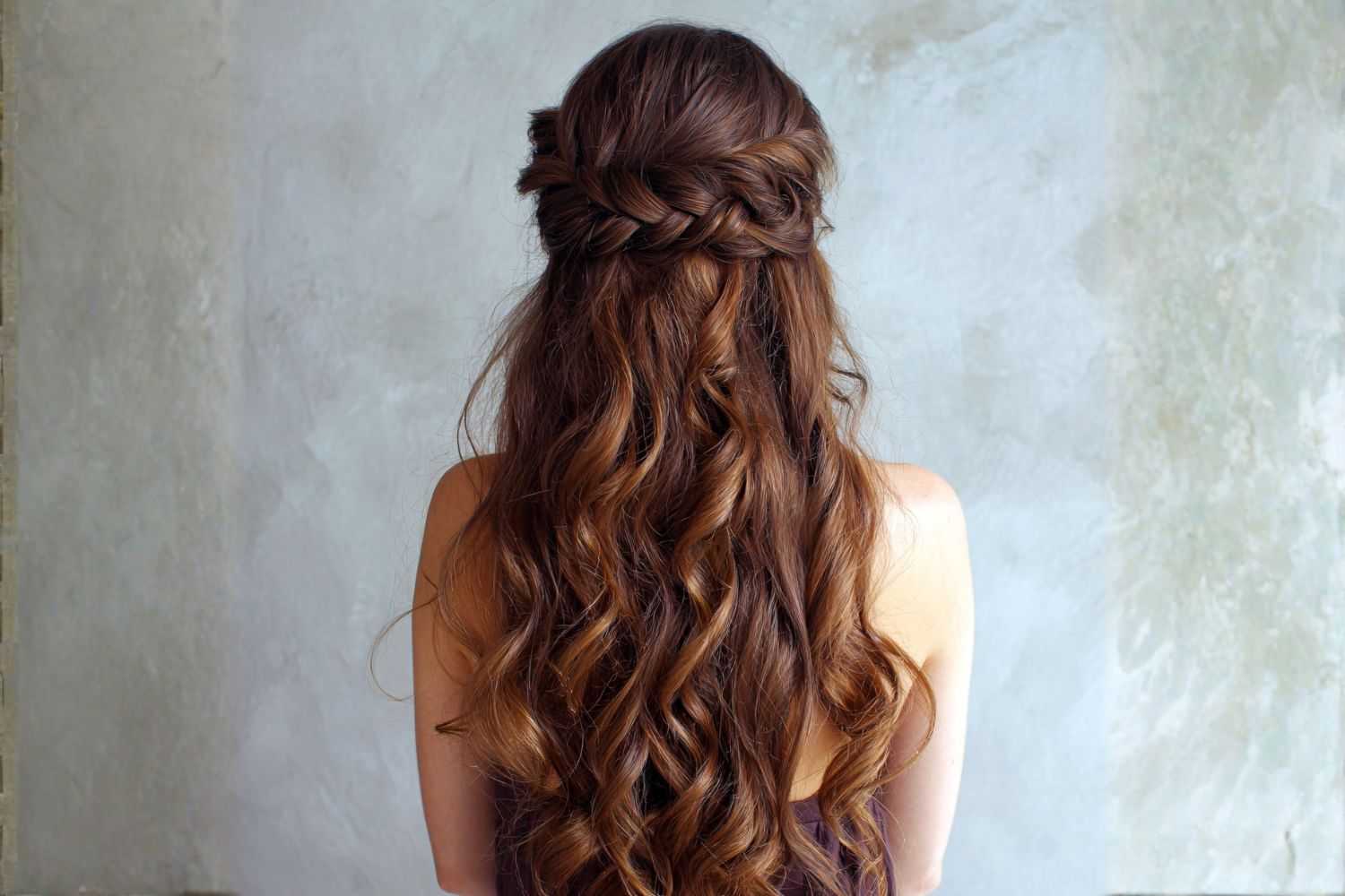Brunette woman with long, wavy hair styled in a braided half updo, facing a textured wall.